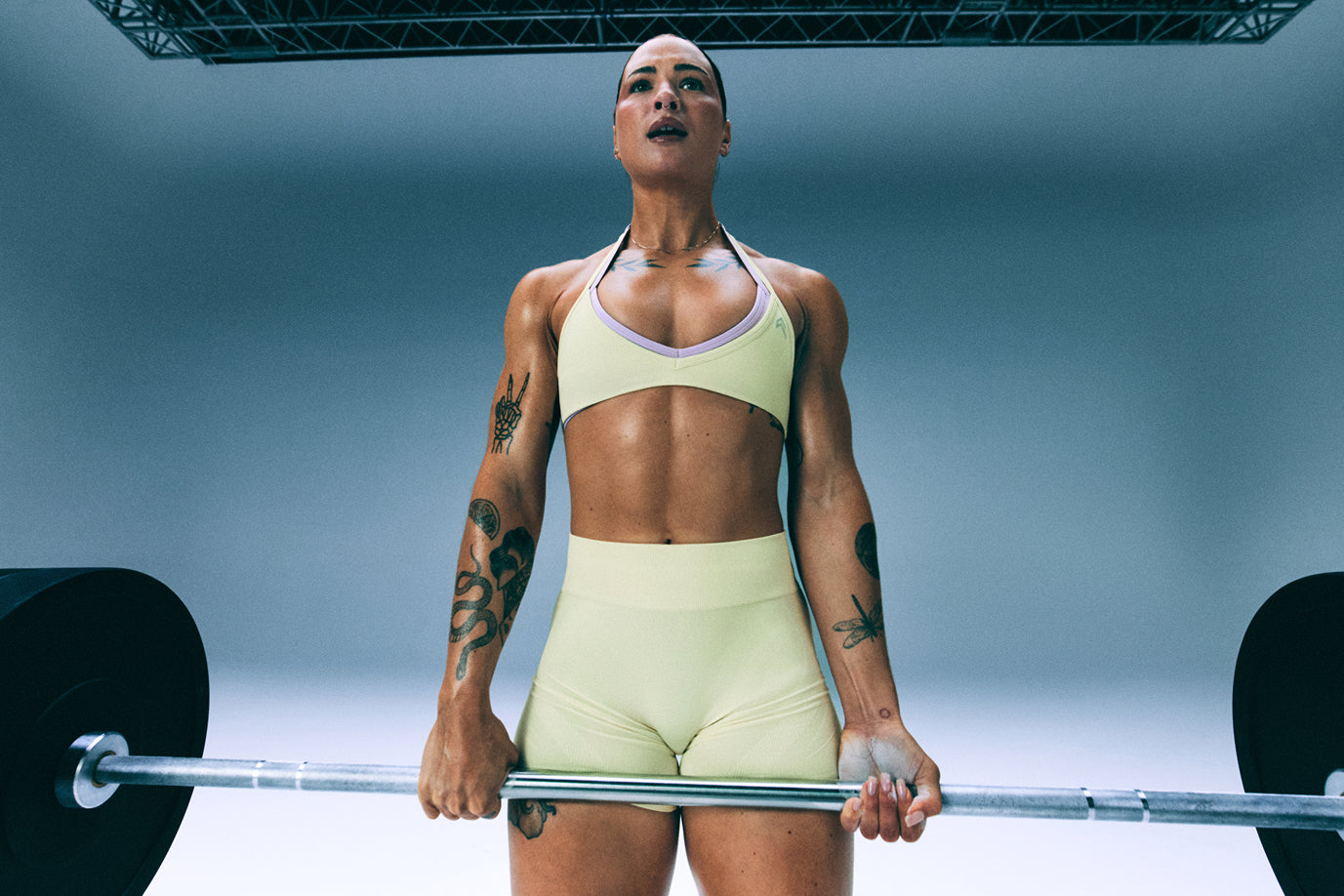 A woman shown from the legs up lifting a barbell and wearing our EnhanceLift™ bralettes layered in Pastel Yellow and Lavender Purple.